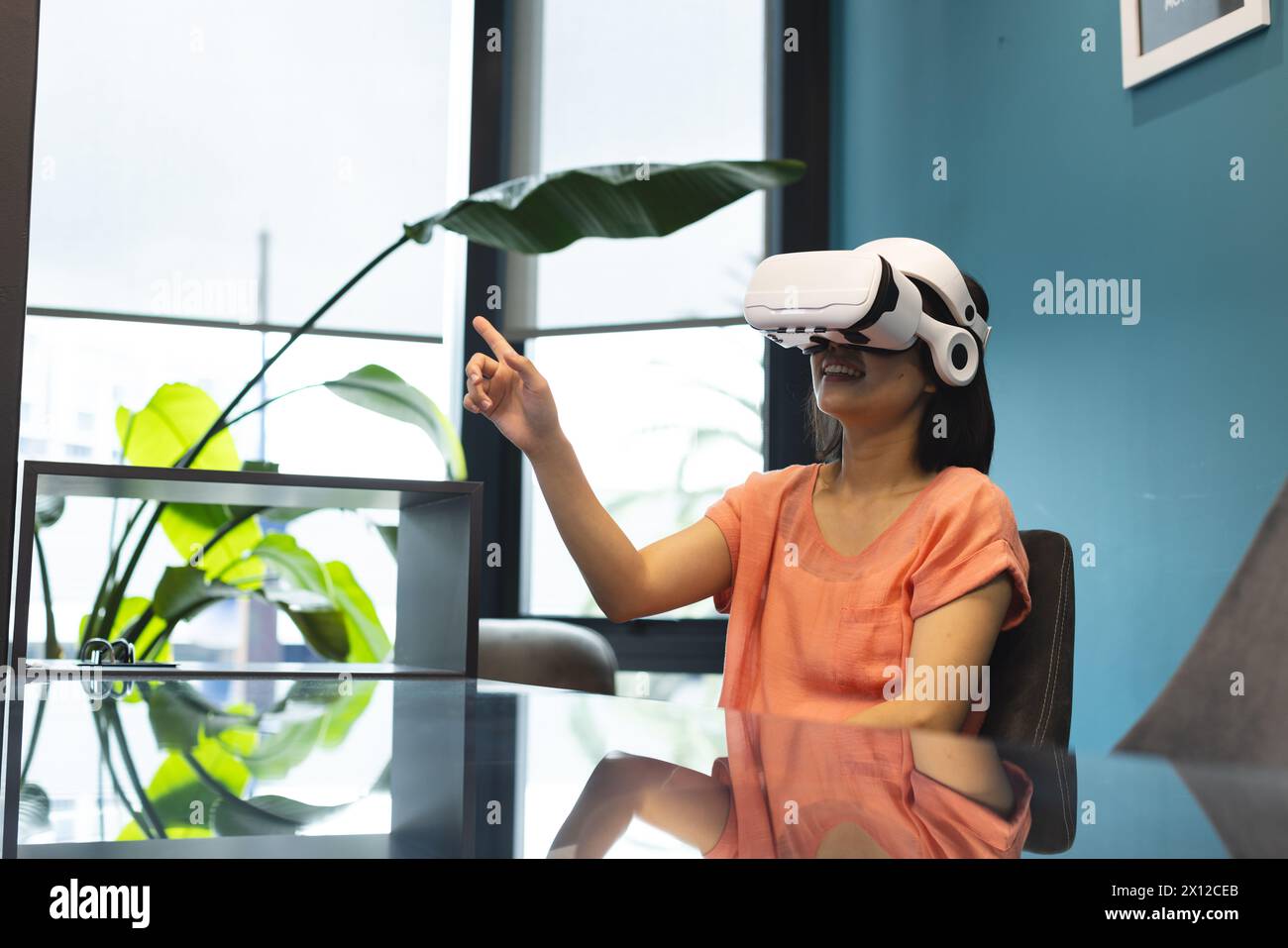 Una giovane donna asiatica che indossa un visore VR sta puntando a qualcosa in un moderno ufficio aziendale Foto Stock