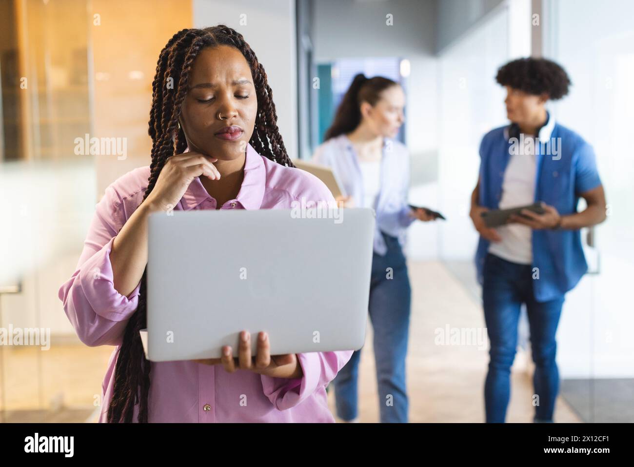 Donna birazziale che tiene un laptop, pensa, colleghi che camminano dietro in un moderno ufficio aziendale Foto Stock