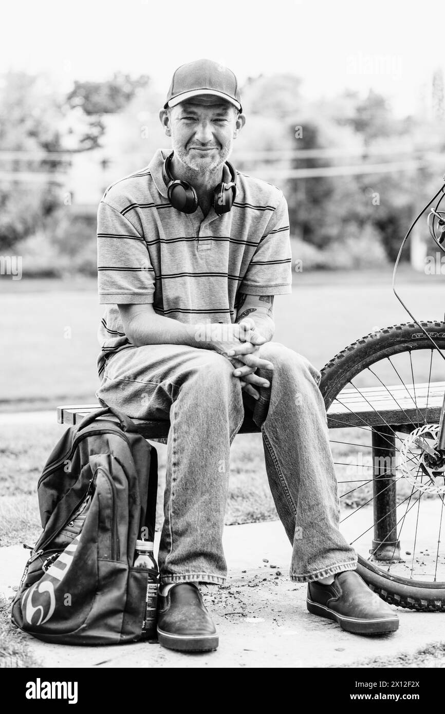 Ubriaco uomo caucasico seduto sulla panchina del parco con zaino e bicicletta Foto Stock