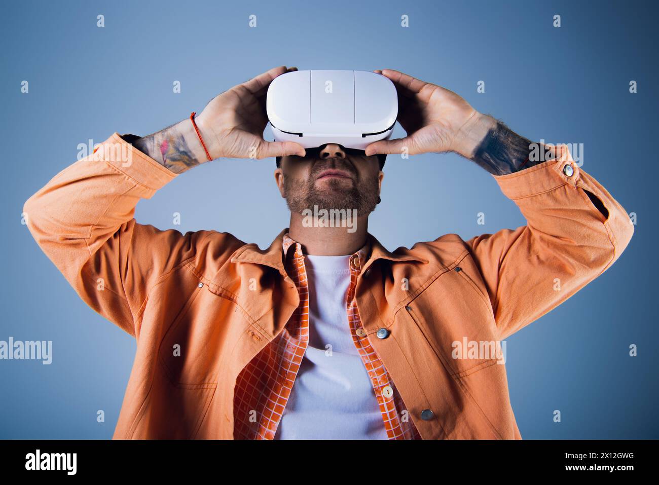 Un uomo con una camicia arancione solleva un visore vr bianco sopra la testa in un ambiente da studio. Foto Stock