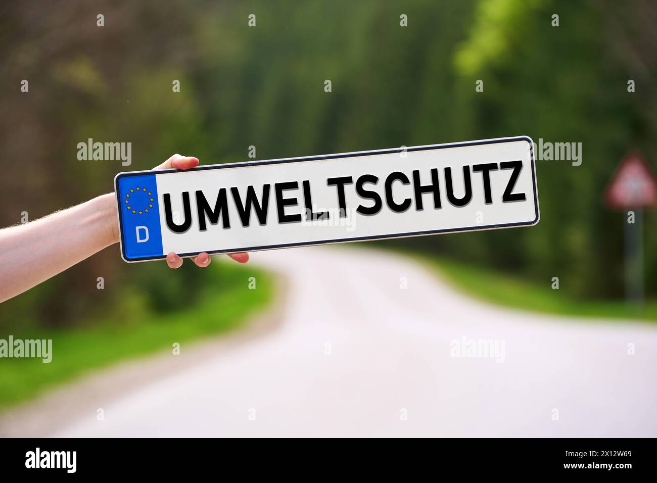 15 aprile 2024: Mano che tiene una targa davanti a una strada di campagna in verde con l'iscrizione: Tutela dell'ambiente. FOTOMONTAGGIO *** mano hält ein Kfz-Kennzeichen vor eine Landstraße in Grüner Natur mit der Aufschrift: Umweltschutz. FOTOMONTAGE Foto Stock
