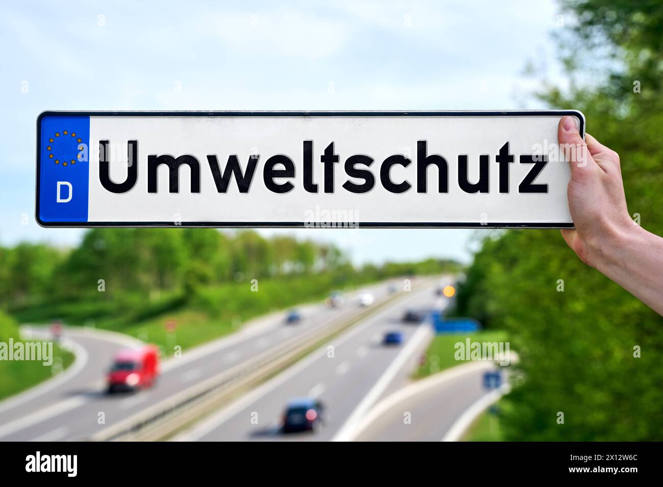 15 aprile 2024: Mano che tiene una targa davanti a un'autostrada con auto in movimento con l'iscrizione: Protezione dell'ambiente. FOTOMONTAGGIO *** mano hält ein Kfz-Kennzeichen vor eine Autobahn mit fahrenden Autos mit der Aufschrift: Umweltschutz. FOTOMONTAGE Foto Stock