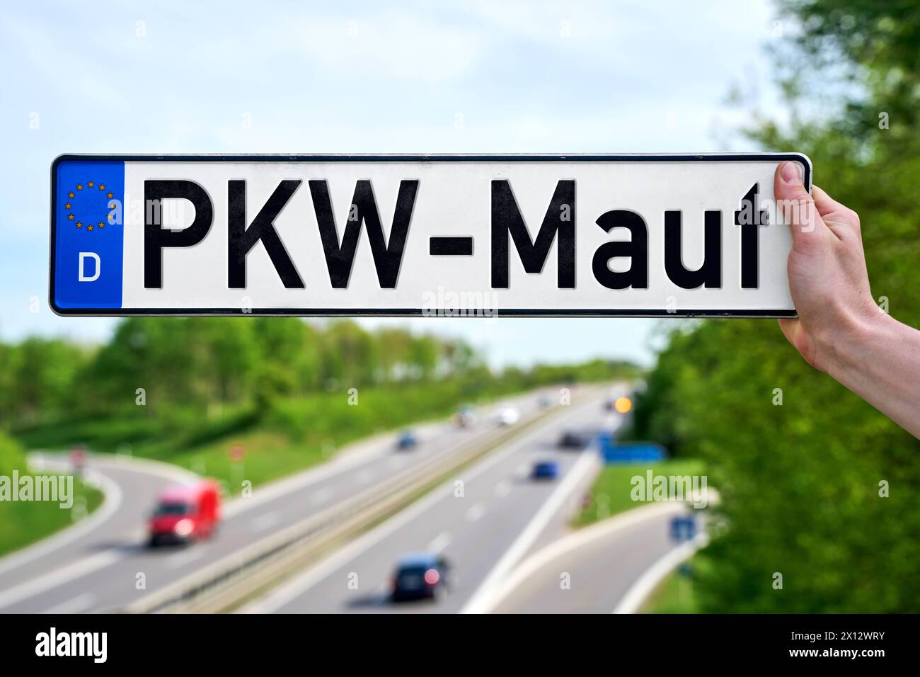 15 aprile 2024: Mano che tiene una targa di fronte a un'autostrada trafficata con l'iscrizione: KFZ-Maut. FOTOMONTAGGIO *** mano hält ein Kfz-Kennzeichen vor eine befahrene Autobahn mit der Aufschrift: KFZ-Maut. FOTOMONTAGE Foto Stock