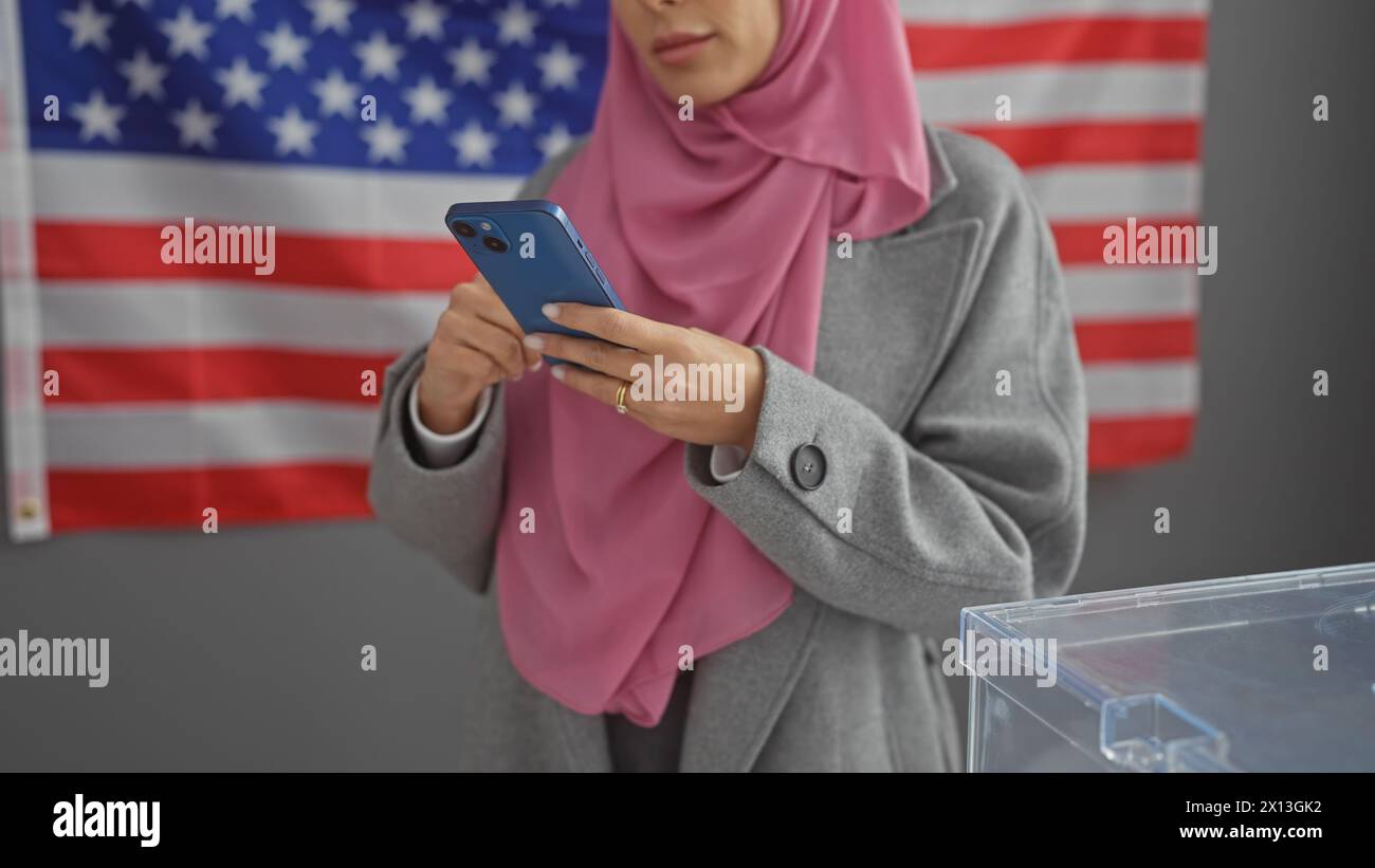 Una giovane donna usa uno smartphone in un centro elettorale degli Stati Uniti con una bandiera americana sullo sfondo. Foto Stock