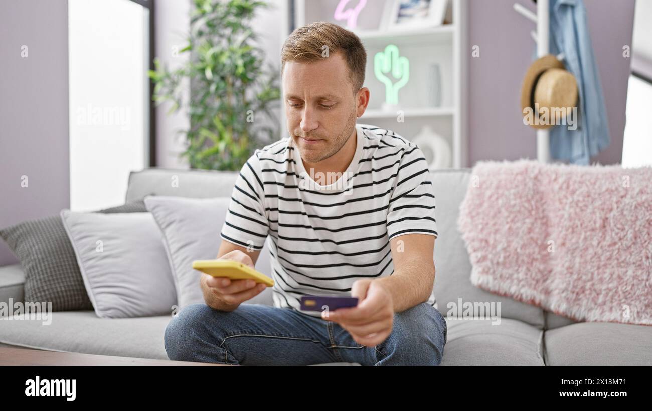 Bellissimo giovane uomo caucasico rilassato, seduto sul divano del salotto, impegnato nello shopping online utilizzando il suo smartphone, scrivendo i dettagli di pagamento con Foto Stock