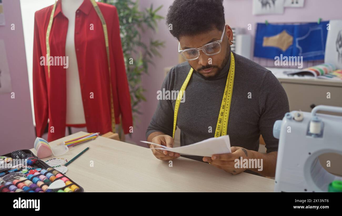 Un sarto afroamericano concentrato che rivede i modelli in un colorato laboratorio di sartoria con accessori da cucire intorno. Foto Stock
