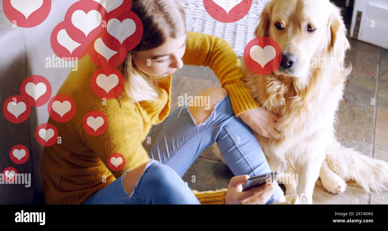 Icone rosse del cuore che fluttuano contro una donna caucasica utilizzando lo smartphone mentre il cane è seduto sul pavimento Foto Stock