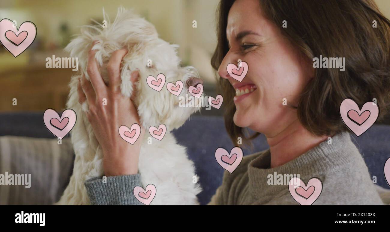 Più icone di cuore rosa fluttuano contro una donna caucasica che bacia il suo cane a casa Foto Stock