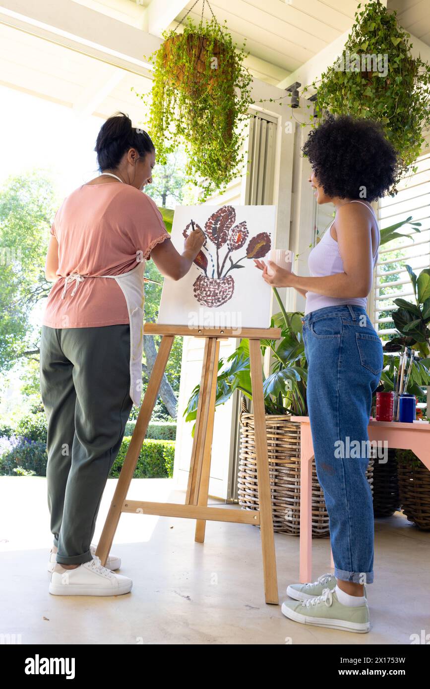 Donna birazziale matura e giovane donna birazziale stanno dipingendo insieme sul portico di casa. Entrambi hanno i capelli ricci, più vecchi nella maglia casual e nei pantaloni, lo younge Foto Stock