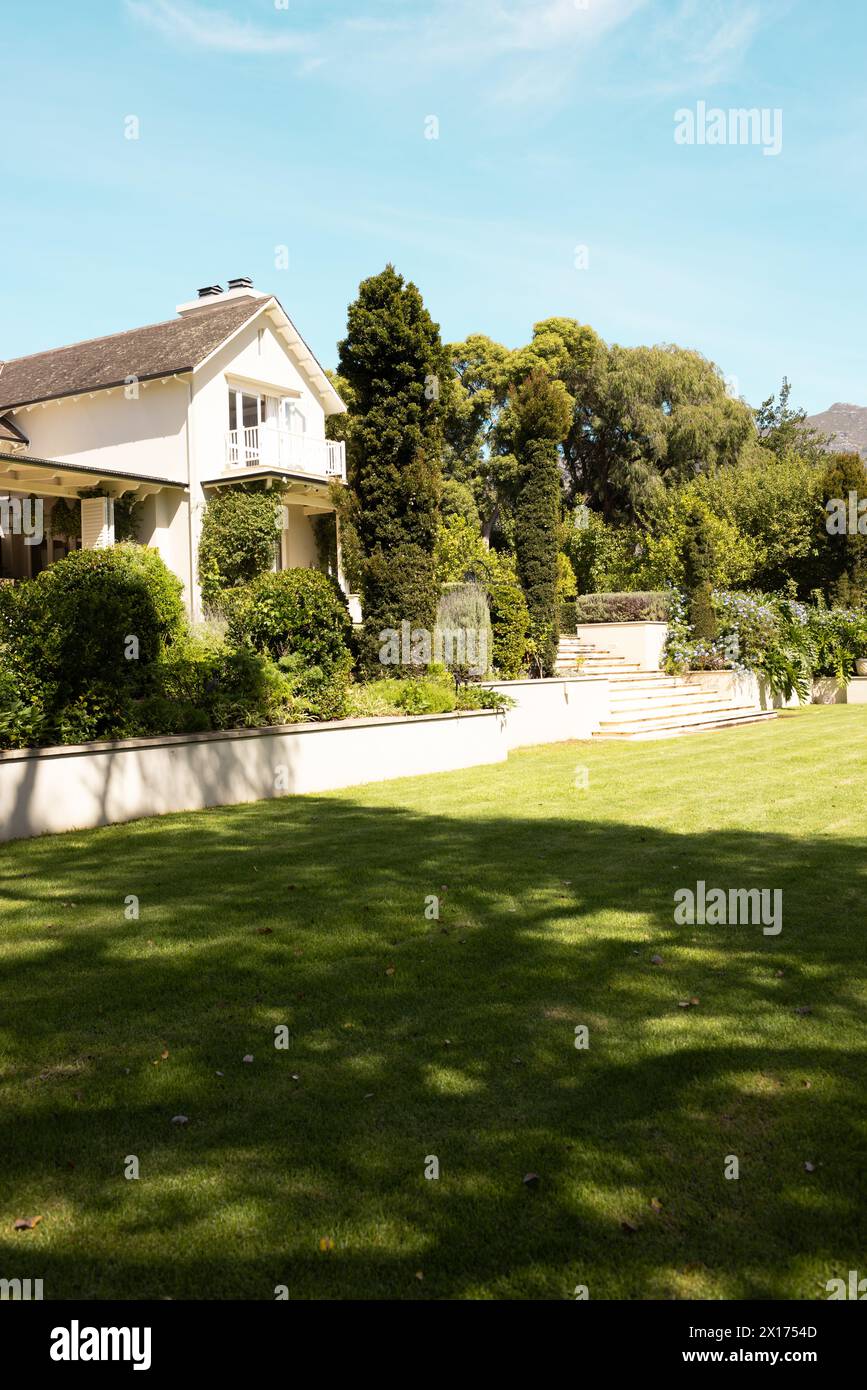 Il lussureggiante prato verde conduce a una grande casa bianca con alberi a casa. La luce solare filtra le foglie, proiettando ombre sull'erba ben tenuta, inalterate. Foto Stock