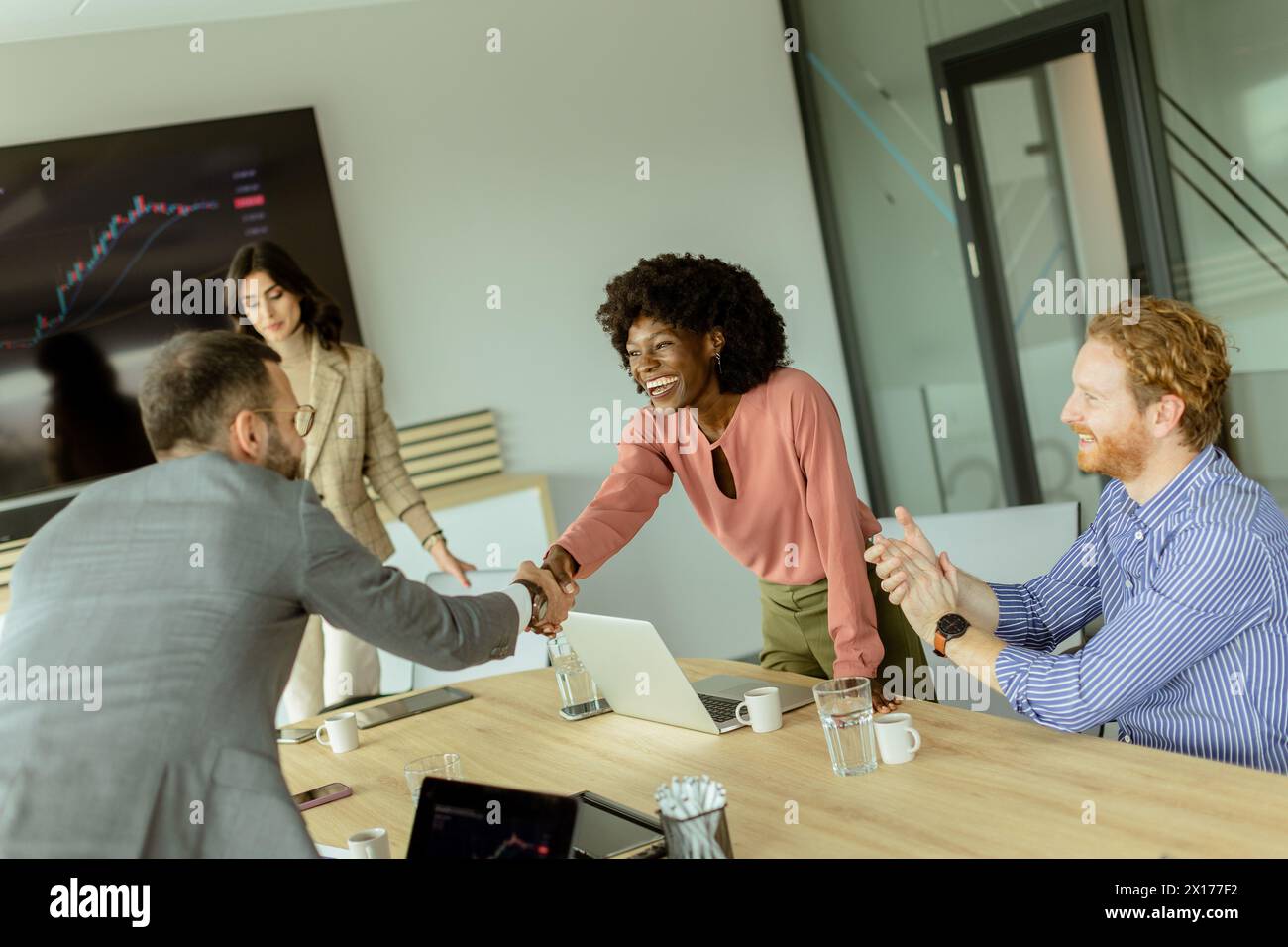 Due colleghi stringono la mano in accordo, mentre un altro paio applaude al loro successo in una sala conferenze ben illuminata Foto Stock