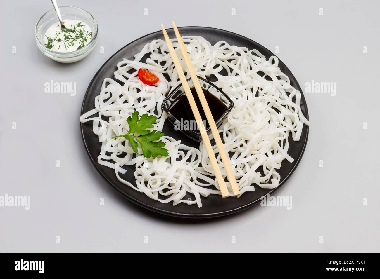 Spaghetti di riso con salsa, bastoncini di bambù sul piatto nero. Pomodori ciliegini nel recipiente con salsa bianca sul tavolo. Sfondo grigio. Vista dall'alto. Foto Stock