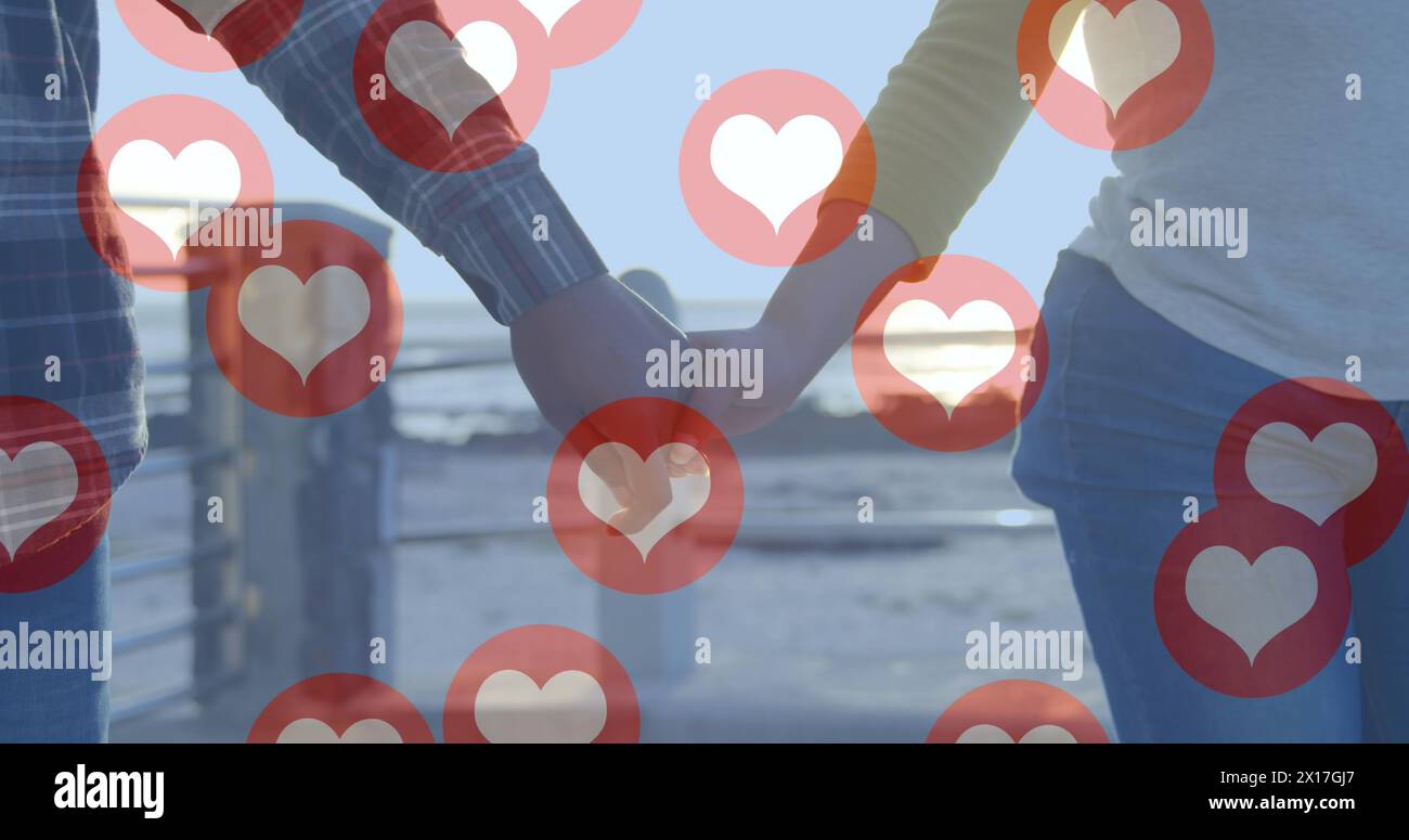 Immagine di icone rosse a forma di cuore che fluttuano sulla sezione centrale di una coppia innamorata che si tiene per mano in riva al mare Foto Stock