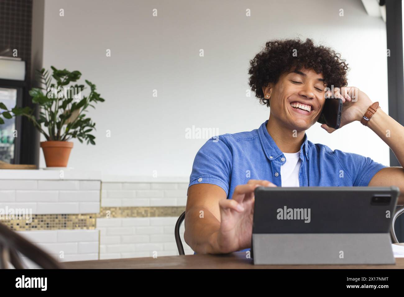 Un giovane uomo birazziale che indossa una camicia blu parla al telefono in un moderno ufficio aziendale. Ha i capelli ricci neri, sta usando una tavoletta, e ha una j Foto Stock