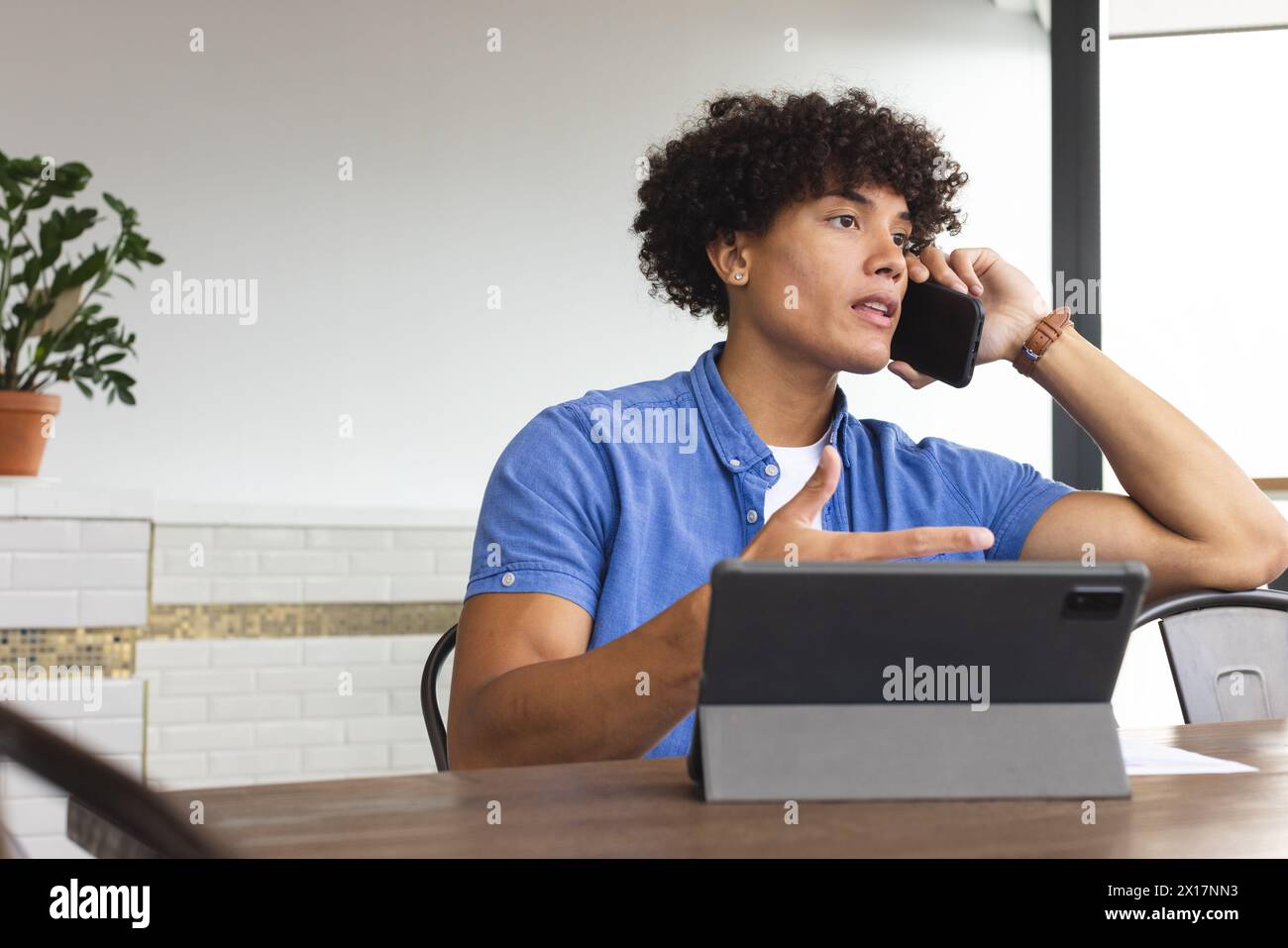 Giovane uomo birazziale che parla al telefono, guarda lontano, seduto a un tavolo in un moderno ufficio d'affari. Ha i capelli neri ricci, indossa una camicia blu, usando Foto Stock