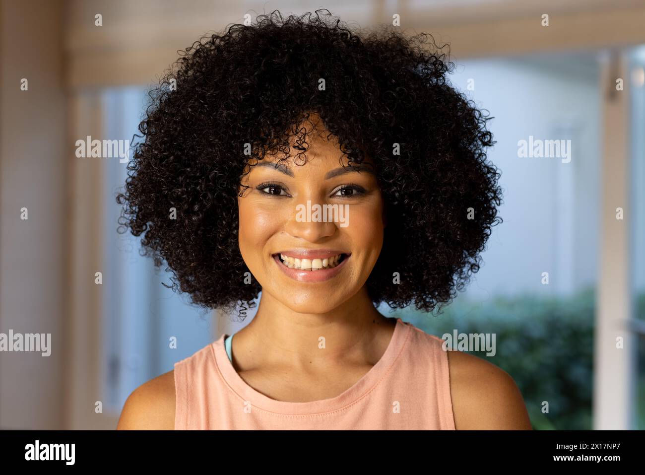 Giovane donna birazziale con capelli neri ricci sorridenti alla macchina fotografica di casa Foto Stock