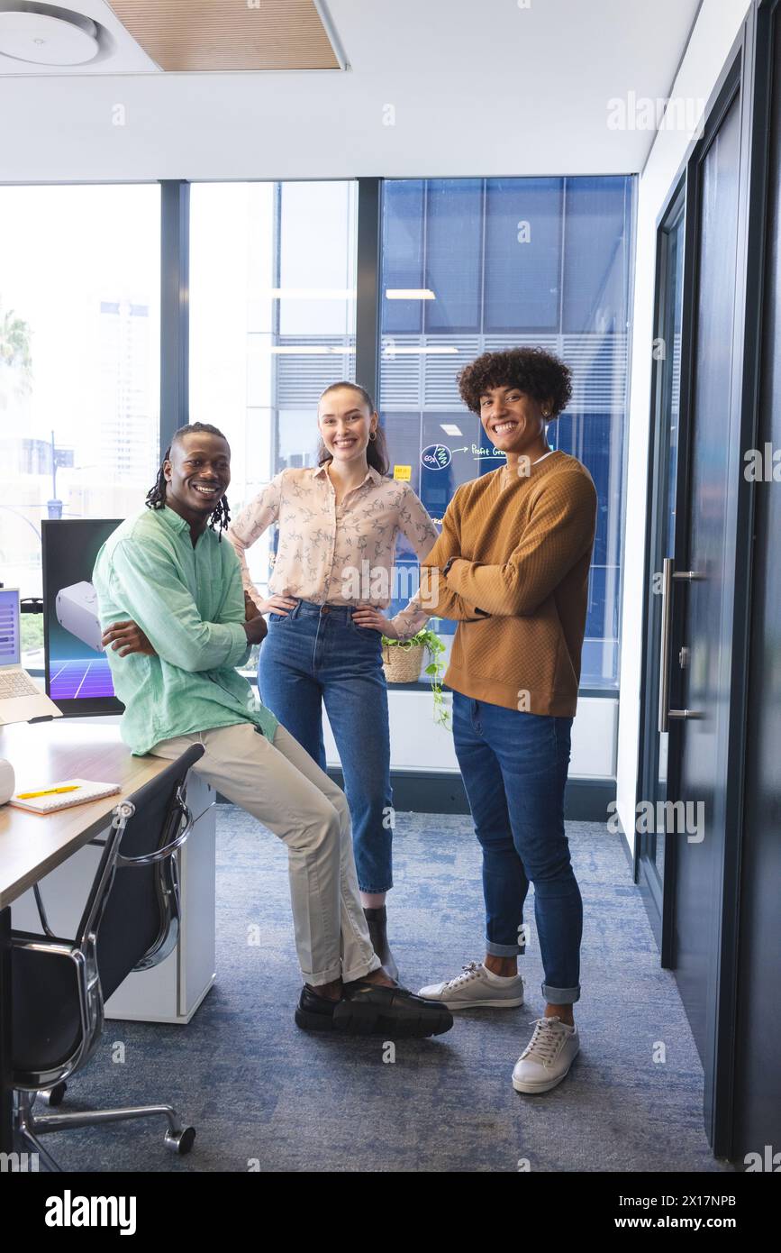 Team diversificato in piedi in ufficio, sorridendo alla telecamera. Uomo afroamericano con camicia verde, donna caucasica con camicetta rosa, uomo birazziale dentro Foto Stock