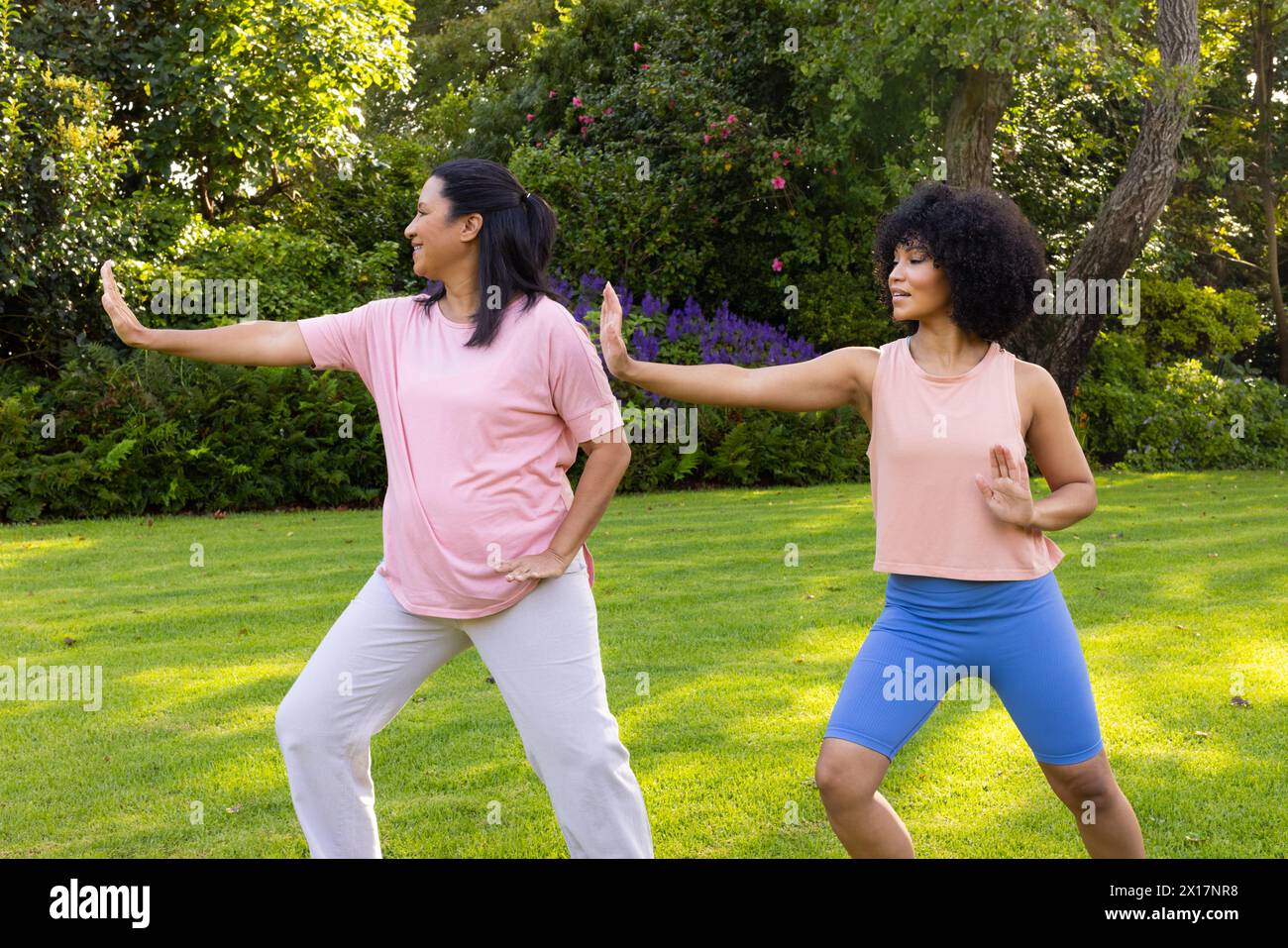 Madre e figlia birazziali che praticano yoga in giardino a casa. Madre che indossa la maglia rosa, figlia in blu, entrambe si divertono in esercizio, inalterate Foto Stock