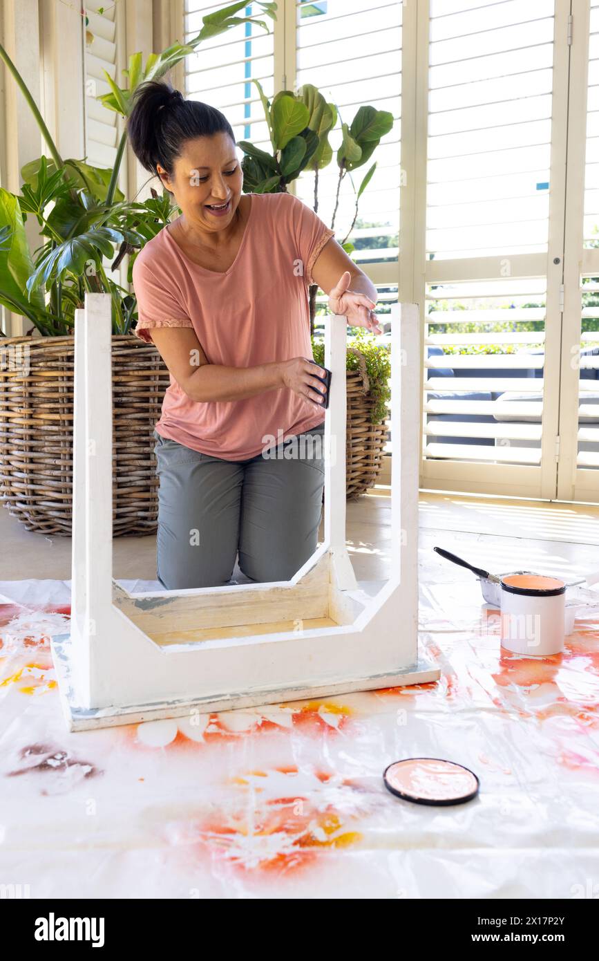 Donna birazziale matura che dipinge mobili a casa in un progetto di upcycling, indossando abiti casual. Ha i capelli scuri, la pelle chiara, e sta usando una vernice Foto Stock