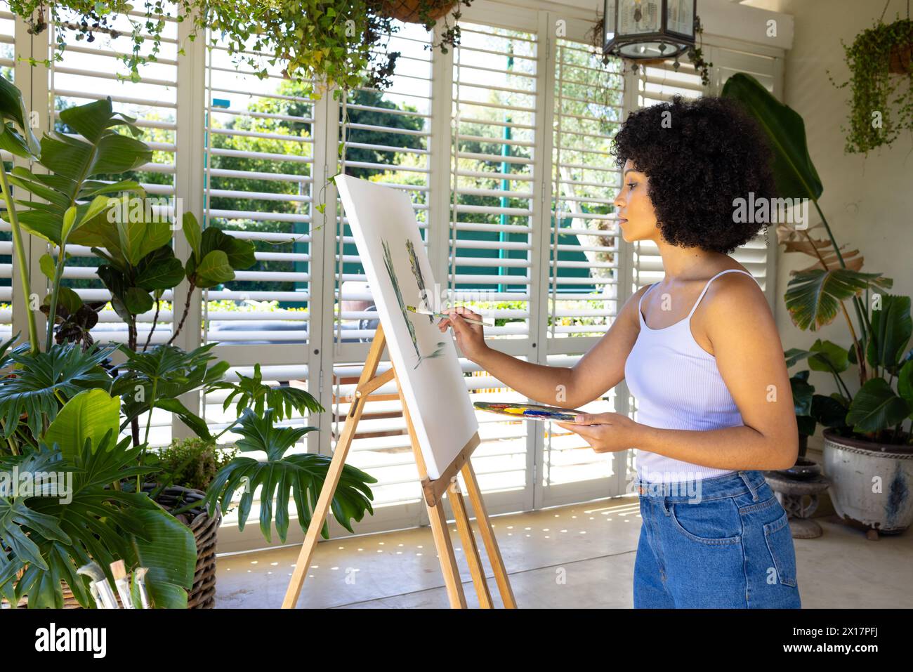 Giovane donna birazziale che dipinge su tela in una stanza piena di piante a casa. Ha i capelli neri ricci, indossa abiti casual, concentrandosi sulla sua arte, inalterata Foto Stock