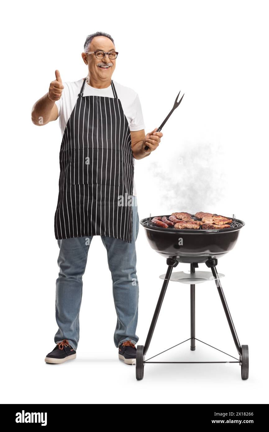 Uomo maturo accanto a una griglia portatile per barbecue che tiene in mano una forchetta e i pollici si sollevano isolati su sfondo bianco Foto Stock