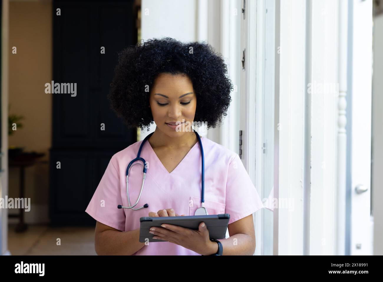 Giovane infermiera birazziale che indossa scrub rosa che controlla la compressa a casa Foto Stock