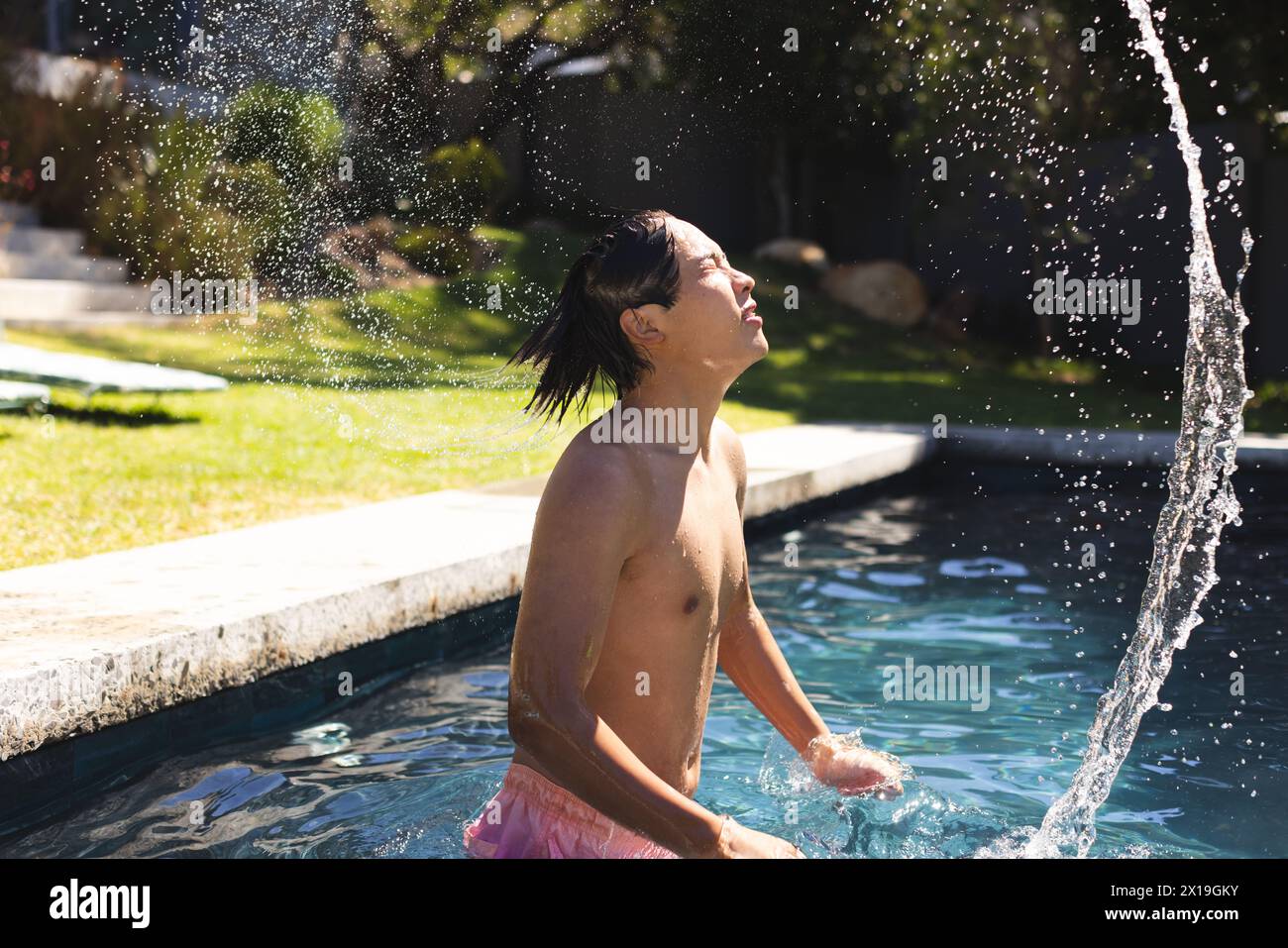 Un adolescente asiatico in piedi in una piscina fuori a casa, spruzzando acqua sul viso Foto Stock