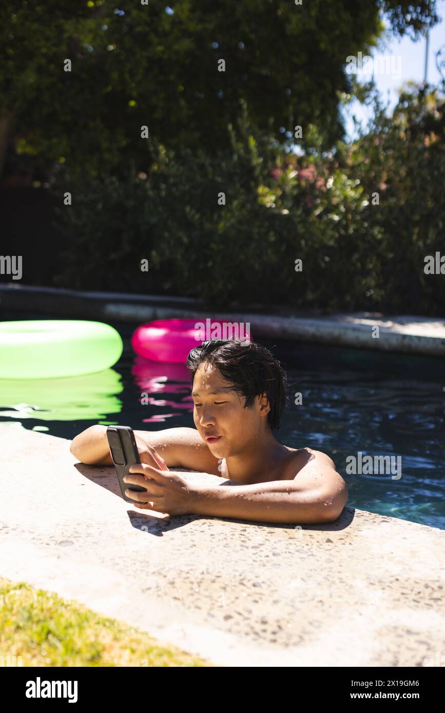 Adolescente asiatico in piscina fuori casa, tenendo lo smartphone, guardando lo schermo, copiando spazio Foto Stock