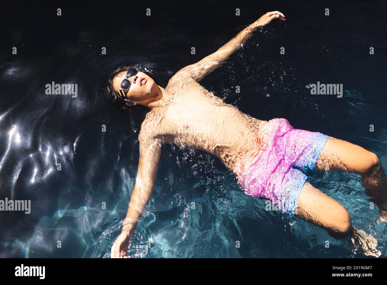 Un adolescente asiatico che indossa occhiali da sole che galleggia nelle acque cristalline della piscina fuori a casa, sembra rilassato Foto Stock