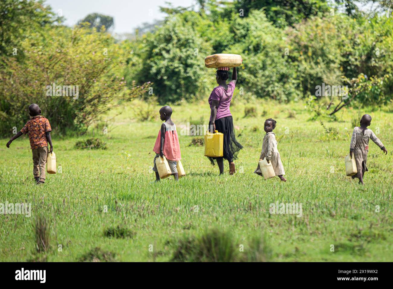 In una foresta africana, una madre ugandese a piedi nudi porta due brocche d'acqua mentre il suo bambino più giovane guarda la macchina fotografica. I bambini, anche a piedi nudi, accompagnano Foto Stock