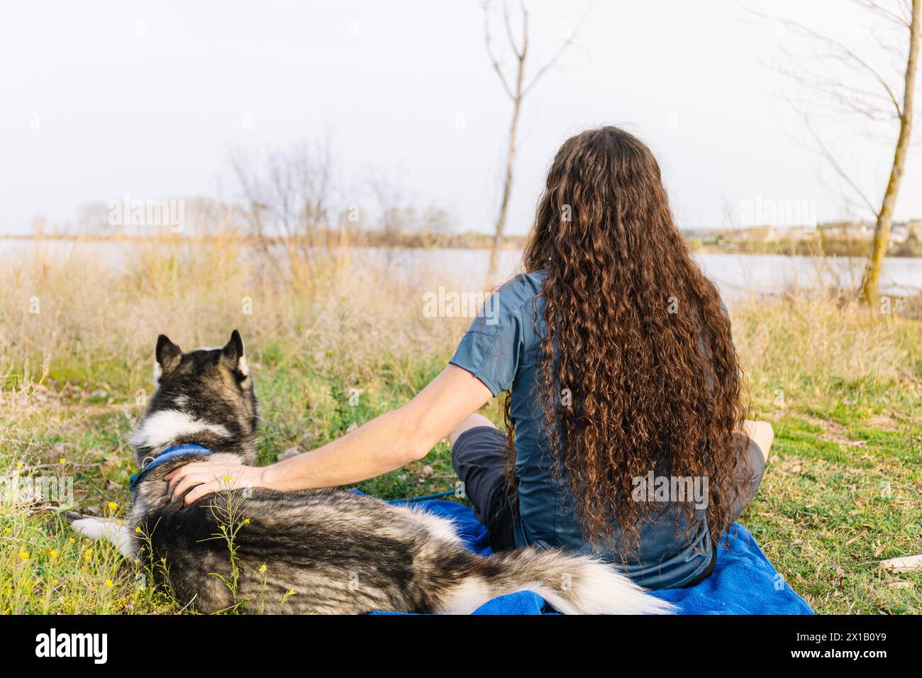 Foto orizzontale un uomo con i capelli ricci si rilassa lungo il fiume, godendosi un momento tranquillo con il suo fedele husky nel morbido abbraccio Foto Stock