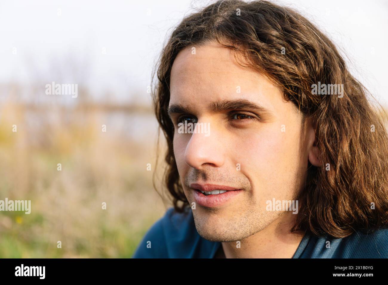 Foto orizzontale un ritratto ravvicinato di un giovane uomo con lunghi capelli ricci, che guarda in lontananza. Concetto di stile di vita. Foto Stock