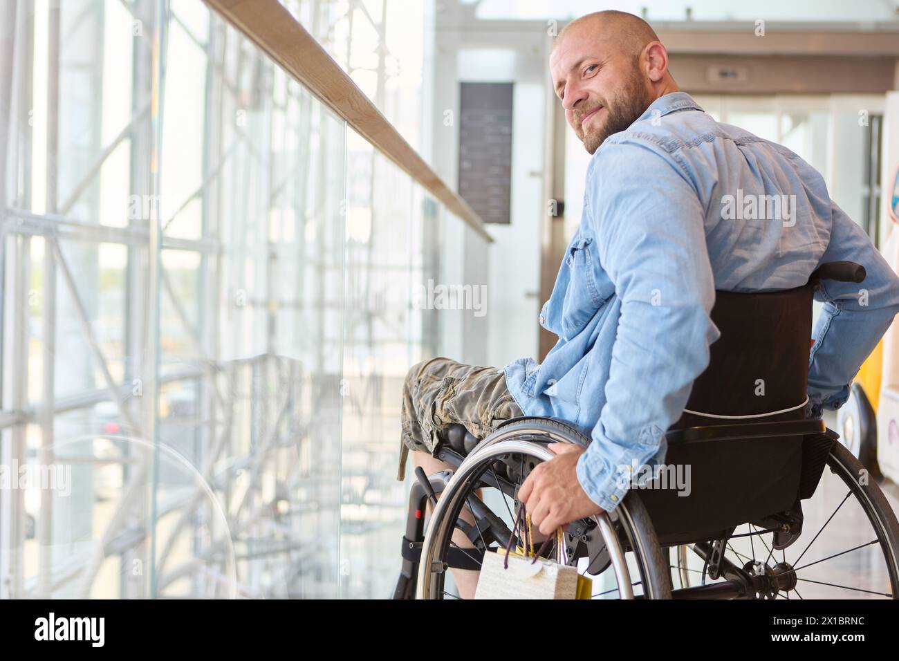 Una persona che utilizza una sedia a rotelle è ritratta sorridendo calorosamente in un edificio moderno e ben illuminato, che mostra un momento di gioia e accessibilità. Foto Stock