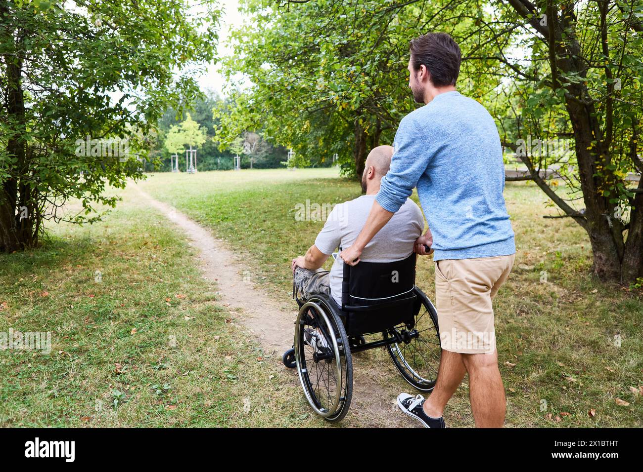 Un momento sereno di amicizia e assistenza in un parco lussureggiante, con un uomo che aiuta un altro che usa una sedia a rotelle a percorrere un sentiero sterrato. Foto Stock