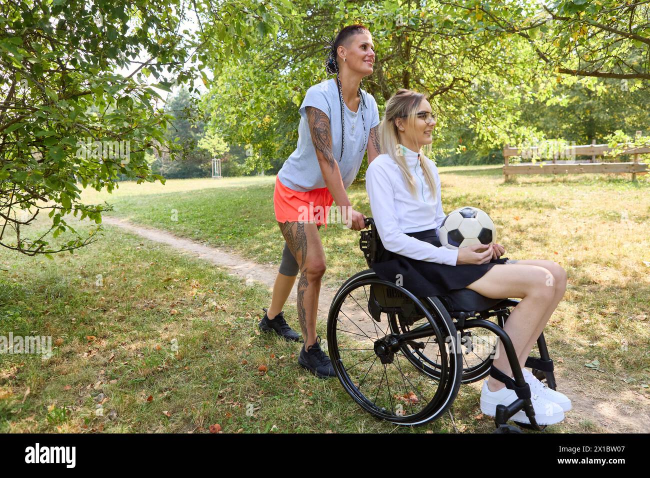 Una donna che usa una sedia a rotelle e un amico in piedi, tiene in mano un pallone da calcio, trascorre una piacevole giornata in un parco verde insieme, incarna amicizia e dentro Foto Stock