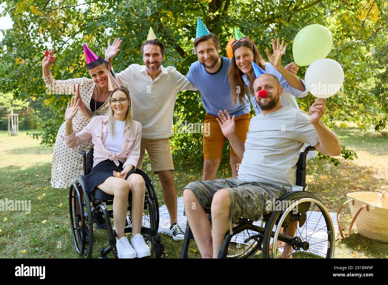 Un gruppo di amici, tra cui persone su sedia a rotelle, partecipa a una vivace festa all'aperto con palloncini, celebrando l'inclusione e la gioia in un lussureggiante set del parco Foto Stock