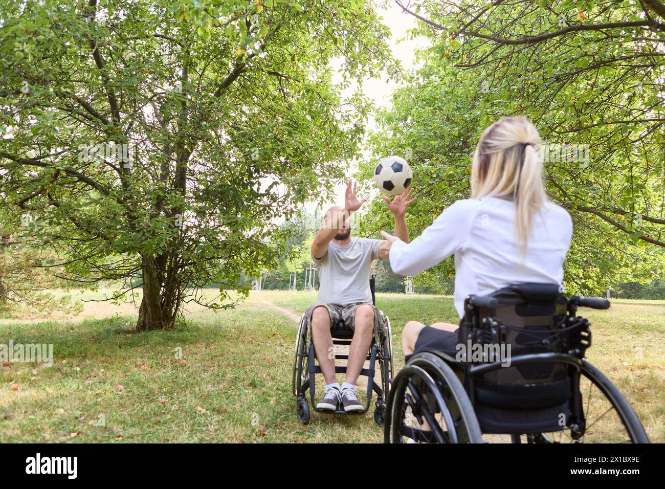 Una scena coinvolgente di inclusività e gioia, che cattura due persone su sedia a rotelle impegnate in una vivace partita di calcio in mezzo al verde lussureggiante in un parco tranquillo. Foto Stock