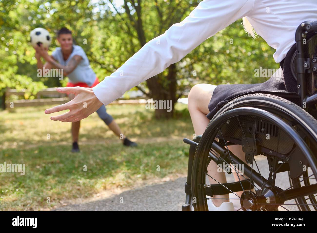 Una scena attiva di una persona in sedia a rotelle che si allontana per giocare a calcio con un amico in un parco lussureggiante e soleggiato, che mostra inclusione e mobilità Foto Stock