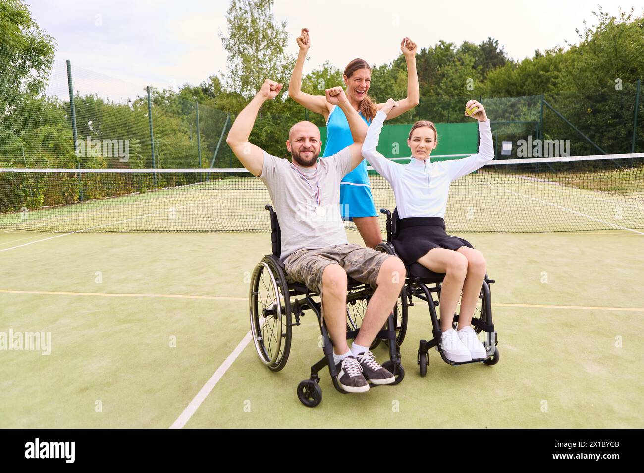 Un allegro gruppo di tre amici, tra cui due persone su sedia a rotelle, che celebrano una vittoria su un campo da tennis all'aperto, mostrando gioia e accessi Foto Stock