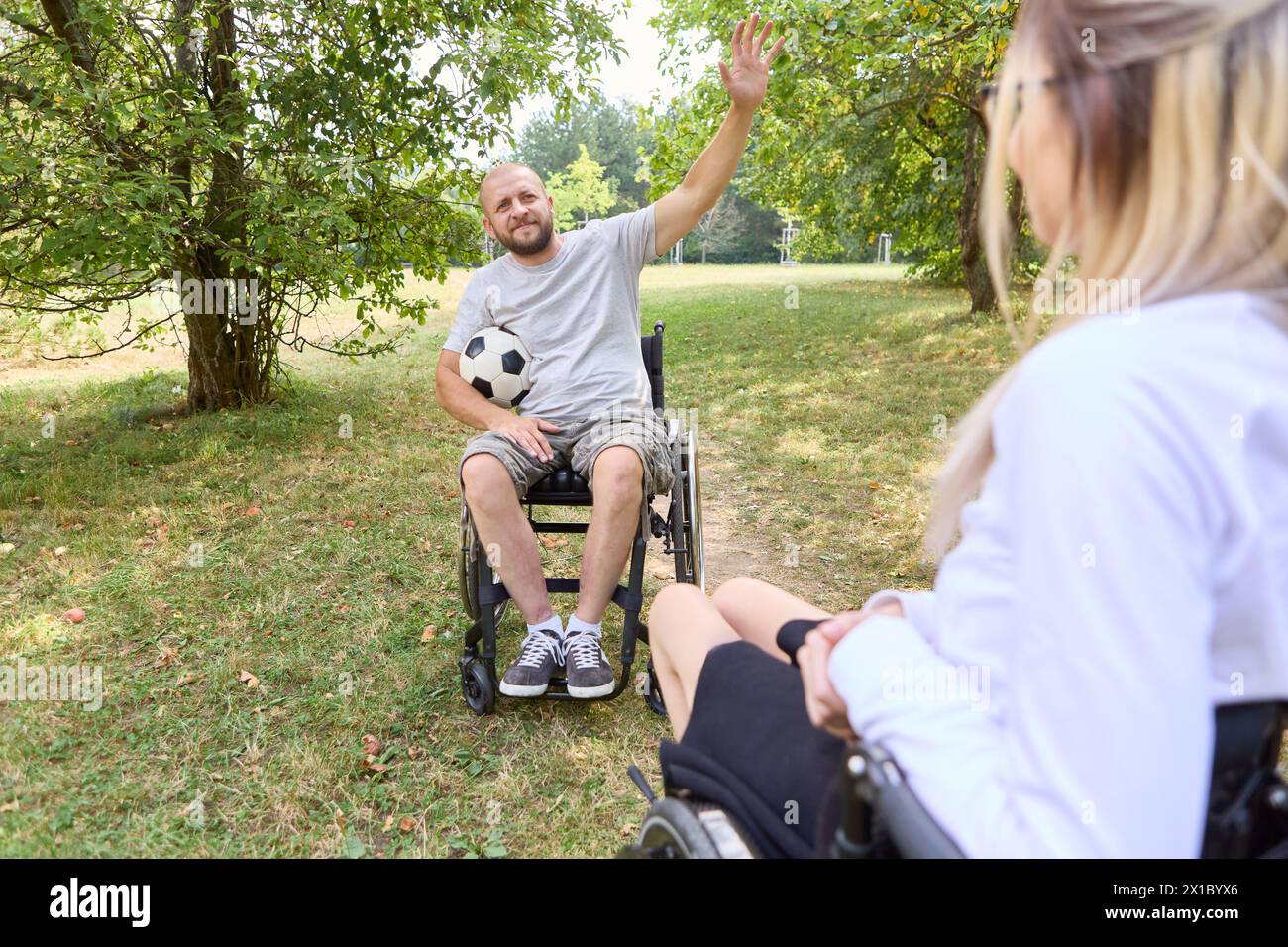 Una scena gioiosa nel parco con una persona che usa una sedia a rotelle con un amico in gamba, mostrando inclusione e cameratismo. Foto Stock