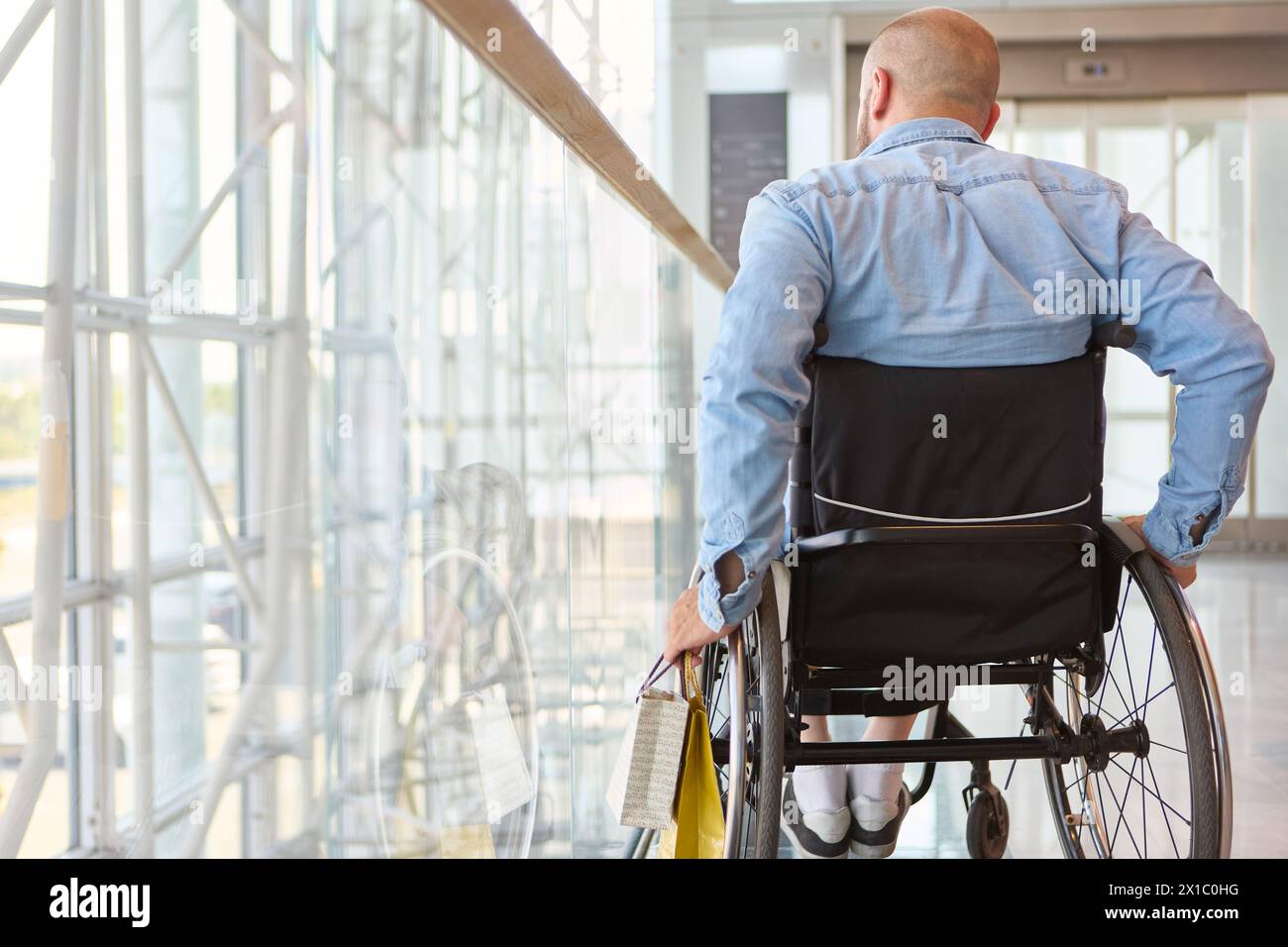 Vista posteriore di una persona su sedia a rotelle che naviga in un edificio moderno, simboleggiando l'accessibilità e l'autosufficienza Foto Stock