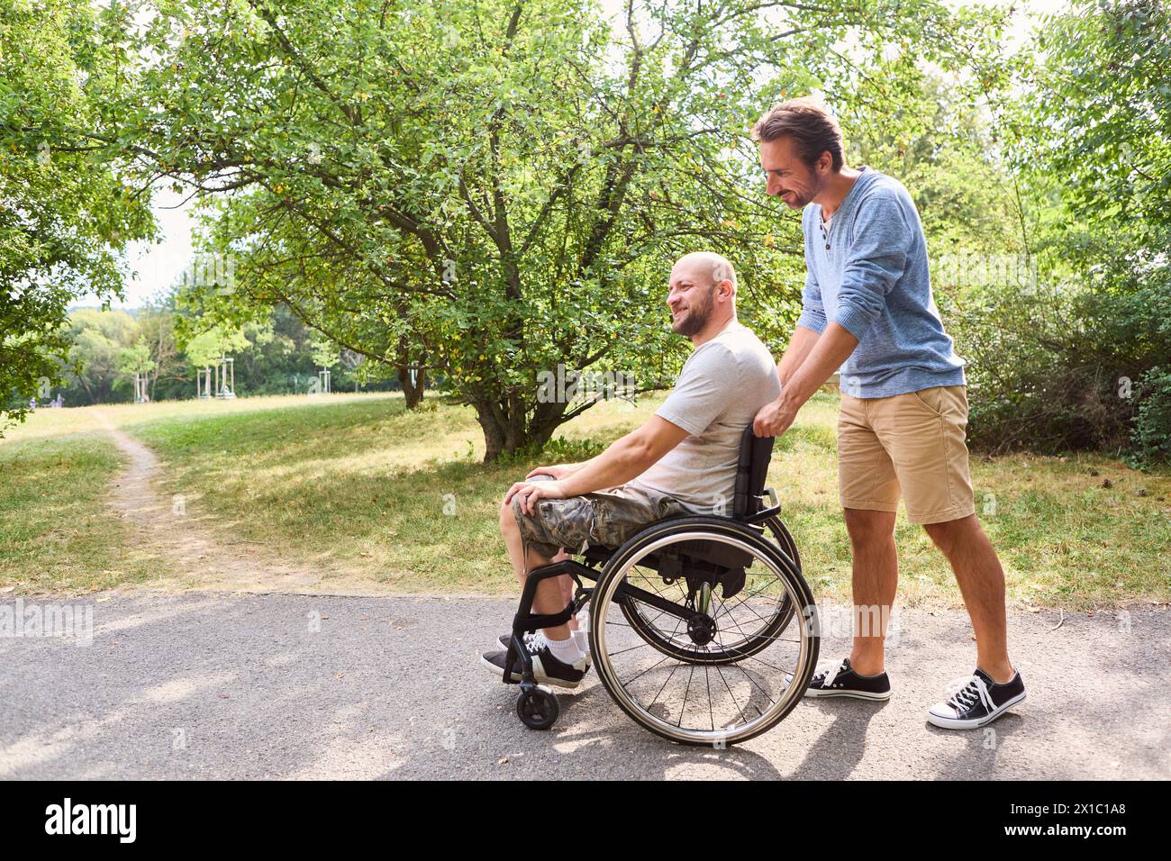 Un uomo su sedia a rotelle che condivide un momento di gioia con il suo amico all'aperto. Si trovano in un parco alberato, che enfatizza l'amicizia e l'inclusione. Foto Stock