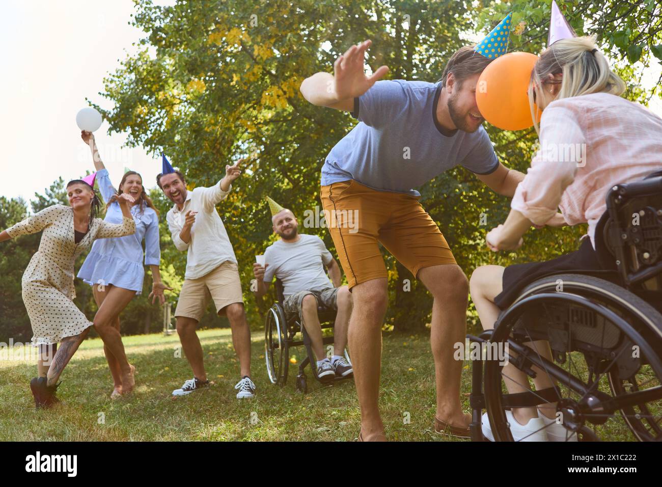 Un gruppo eterogeneo di amici, tra cui una persona su sedia a rotelle, festeggia all'aperto con gioia e cappelli da festa. Foto Stock