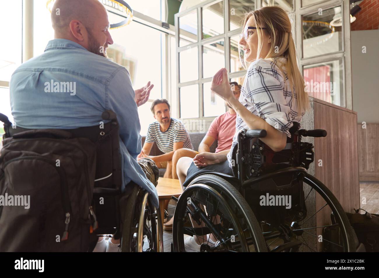 Un gruppo eterogeneo di persone, tra cui persone su sedia a rotelle, che conversano in uno spazio interno ben illuminato, mostrando inclusione e accesso facilitato Foto Stock
