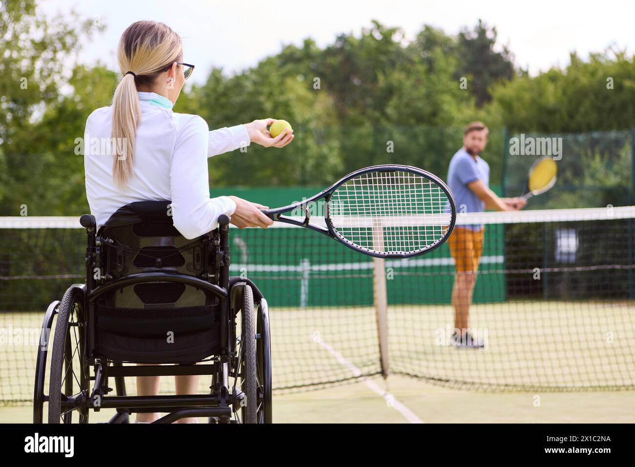 Una donna attiva su una sedia a rotelle che si prepara a giocare a tennis con un partner in piedi su un campo all'aperto che mostra l'inclusione e l'adattabilità negli sport. Foto Stock