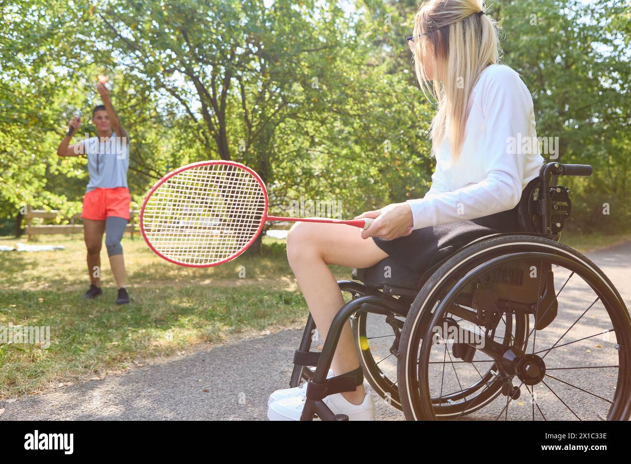 Una donna che usa una sedia a rotelle e la sua amica che gioca a badminton in un parco, raffigurando gioia e stile di vita attivo nonostante le sfide fisiche. Foto Stock