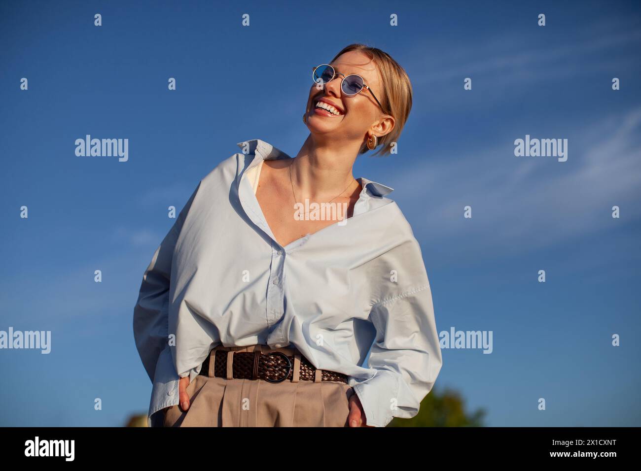 Donna contemporanea con camicia oversize e occhiali da sole, che sorride con gioia sul cielo blu. Atmosfera estiva alla moda, felice ed elegante. Foto Stock