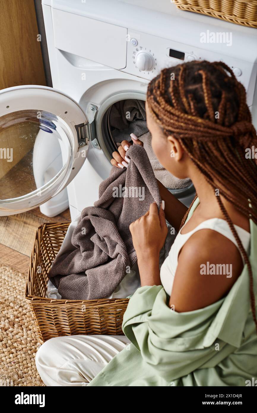 Donna afro-americana con trecce afro sedeva accanto a una lavatrice, facendo il bucato in un bagno. Foto Stock