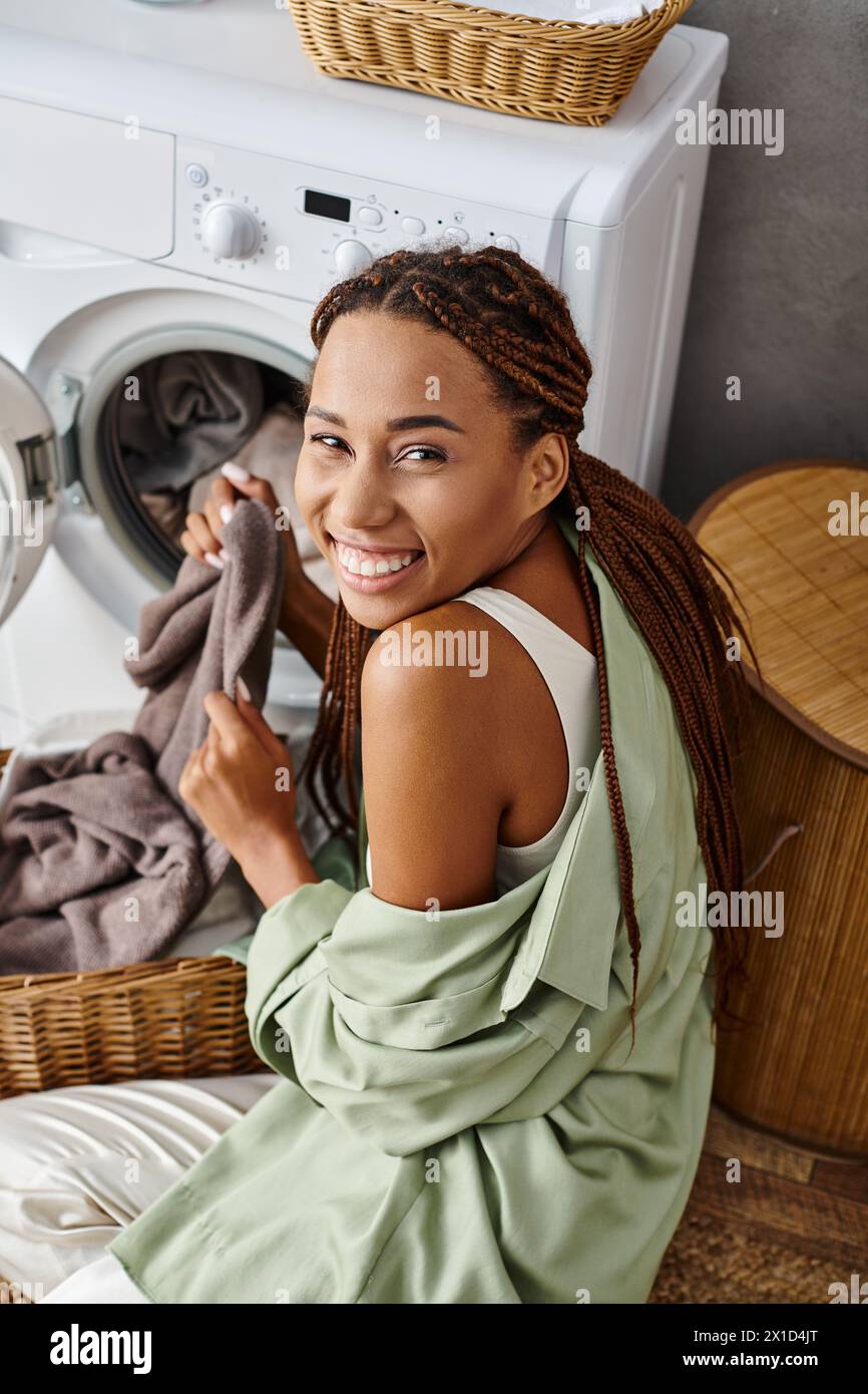 Una donna afroamericana con trecce afro siede sul pavimento accanto a una lavatrice, facendo il bucato in bagno. Foto Stock