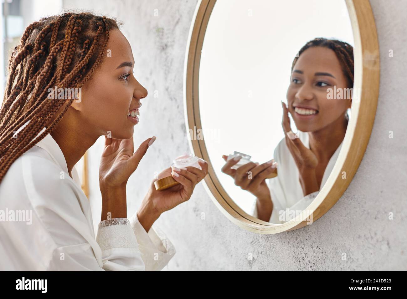 Una donna afroamericana con trecce afro che applica crema in un bagno moderno mentre indossa un accappatoio. Foto Stock